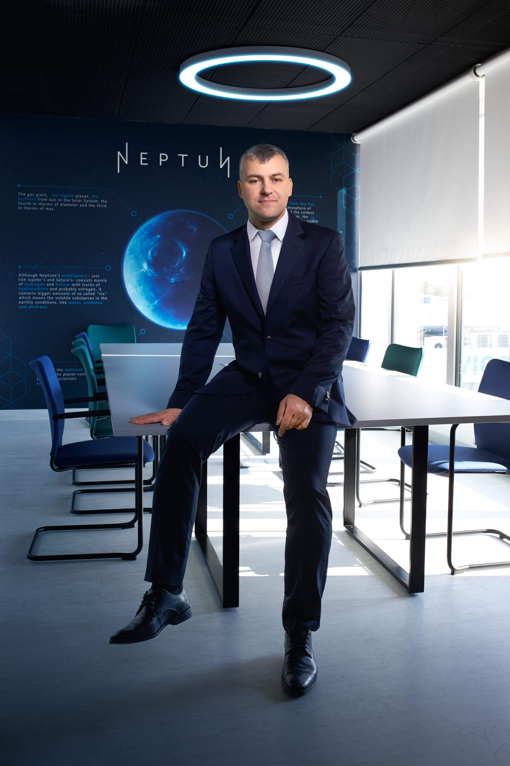 Adam Piotrowski, President of the Board at VIGO Photonics, in a suit sits on a conference table in a modern office with a space-themed background featuring a large image of a planet and the word "NEPTUN." Halo-like ceiling light above. Various chairs are positioned around the table.
