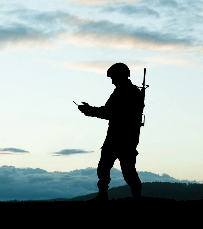 Silhouette of a soldier standing outdoors at dusk, holding a device in one hand and with a rifle slung across their back. Clouds and sky are visible in the background.