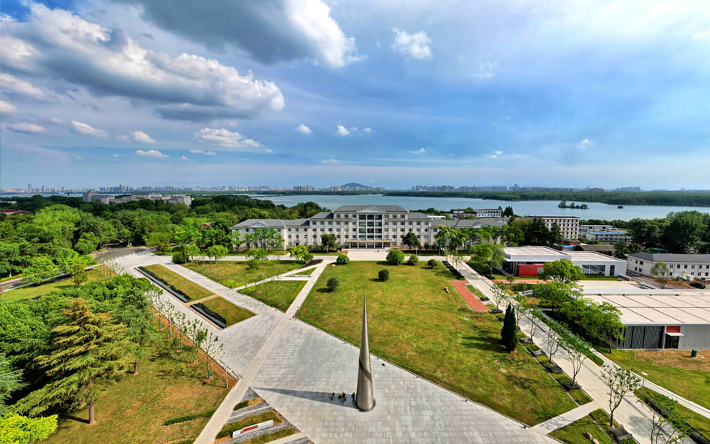Aerial view of a university campus with a central green lawn and paved pathways. Surrounding trees frame a large building. The background features a lake and a distant city skyline under a cloudy sky.