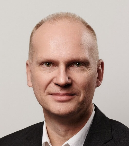 Portrait of a smiling man with a shaved head, wearing a dark suit and white shirt, against a plain background.
