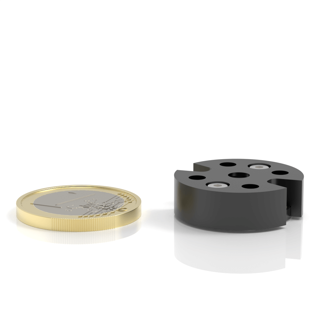 A gold-colored coin lies flat next to a small, round, black cylindrical object with holes and screws, both on a white surface with reflections.