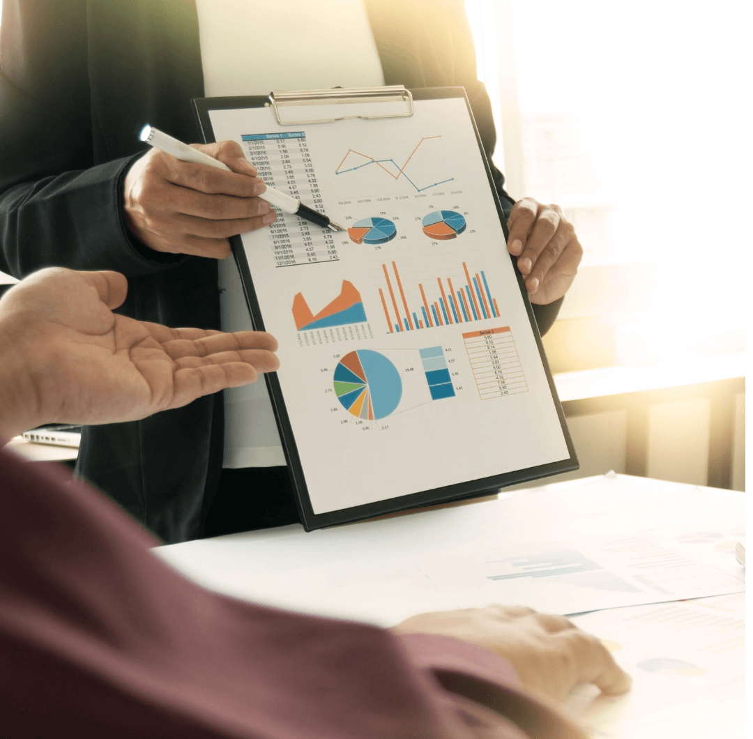 Two people discuss colorful financial charts and graphs on a clipboard in a bright office setting. One person is pointing at the clipboard with a pen while the other listens and gestures with their hand.