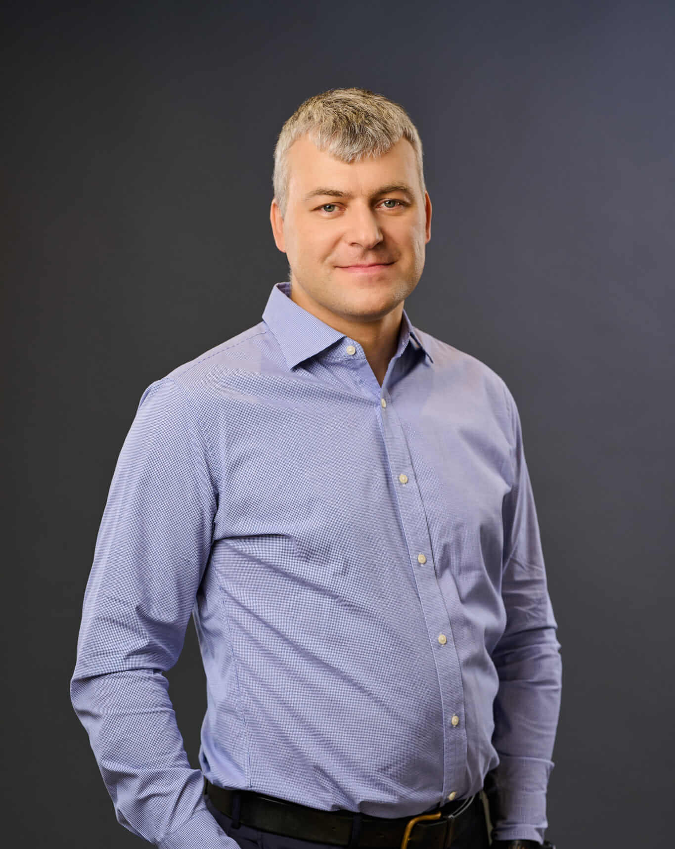 A man with short gray hair wearing a light blue button-up shirt and black pants stands against a plain dark background, smiling with one hand in his pocket.
