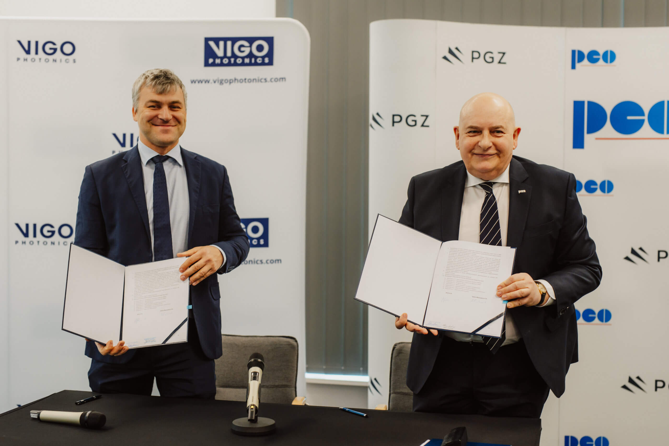 Two men in suits smile and hold up signed documents at a table with microphones. Behind them are banners displaying the logos of VIGO Photonics, PGZ, and PCO, suggesting a formal agreement or partnership.