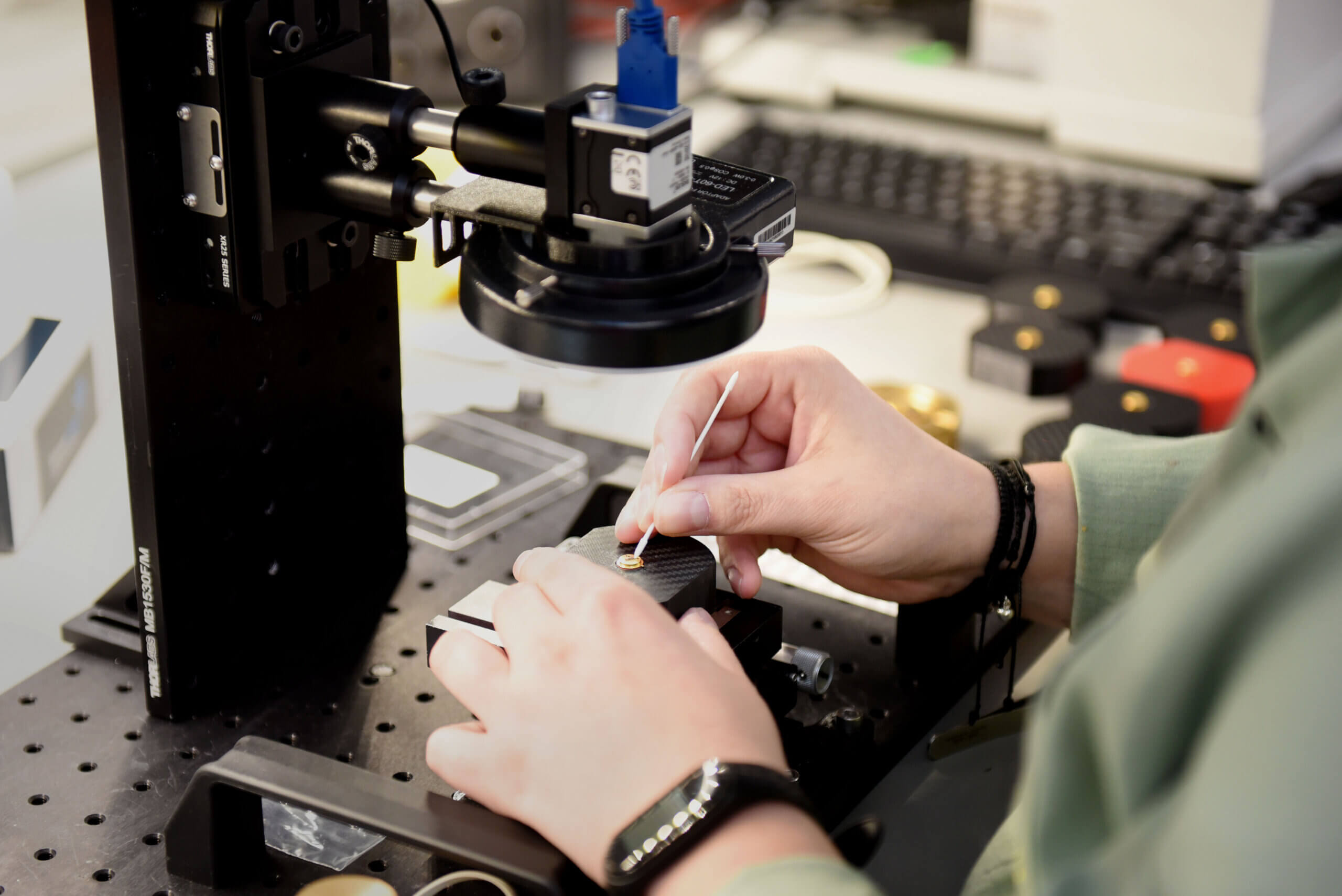 A person uses tweezers to work with small electronic components under a microscope on a lab workstation, with a keyboard and various lab equipment in the background.