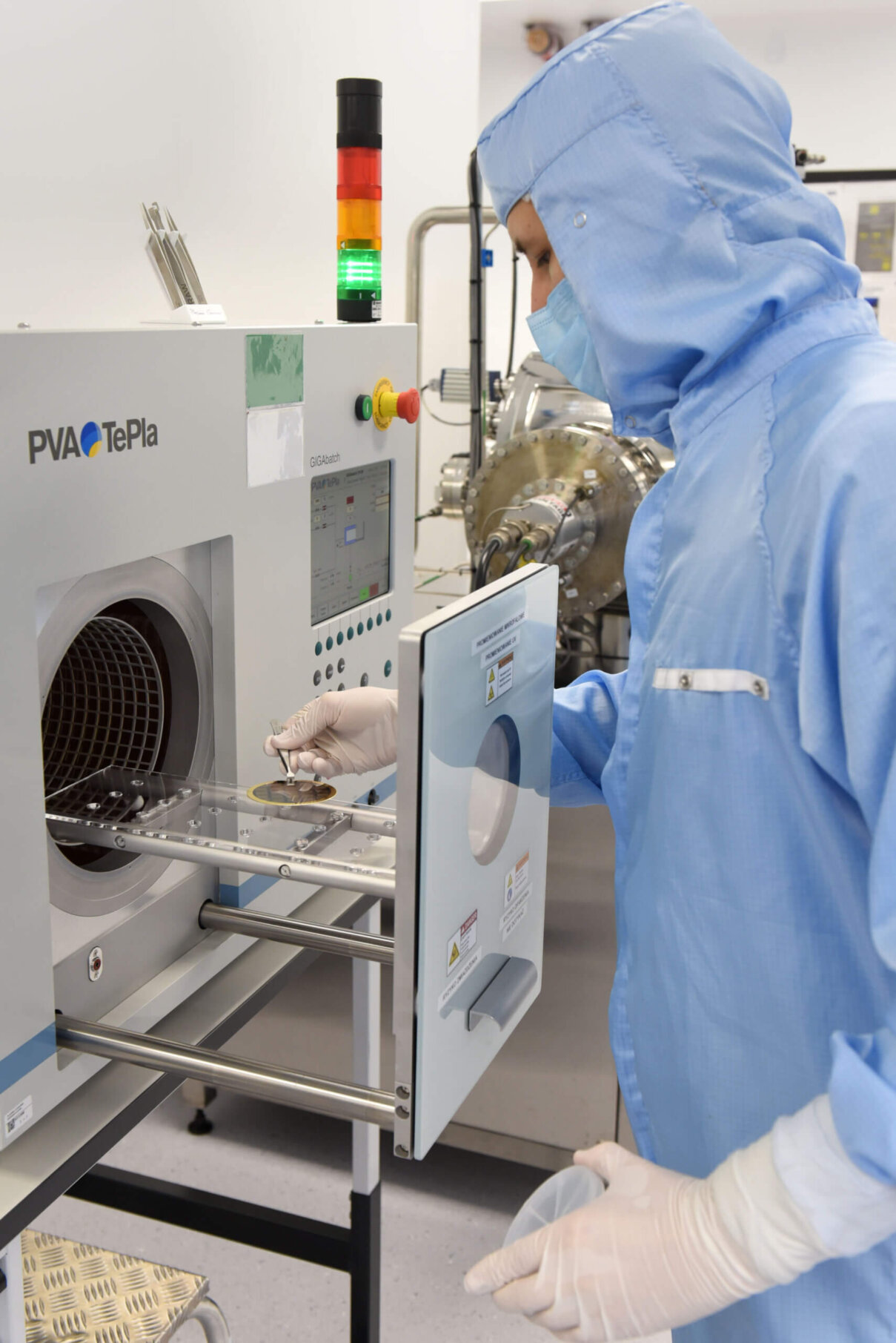 A person in a blue cleanroom suit and gloves operates a PVA TePla machine, placing items inside. The environment appears to be a laboratory with scientific equipment visible in the background.