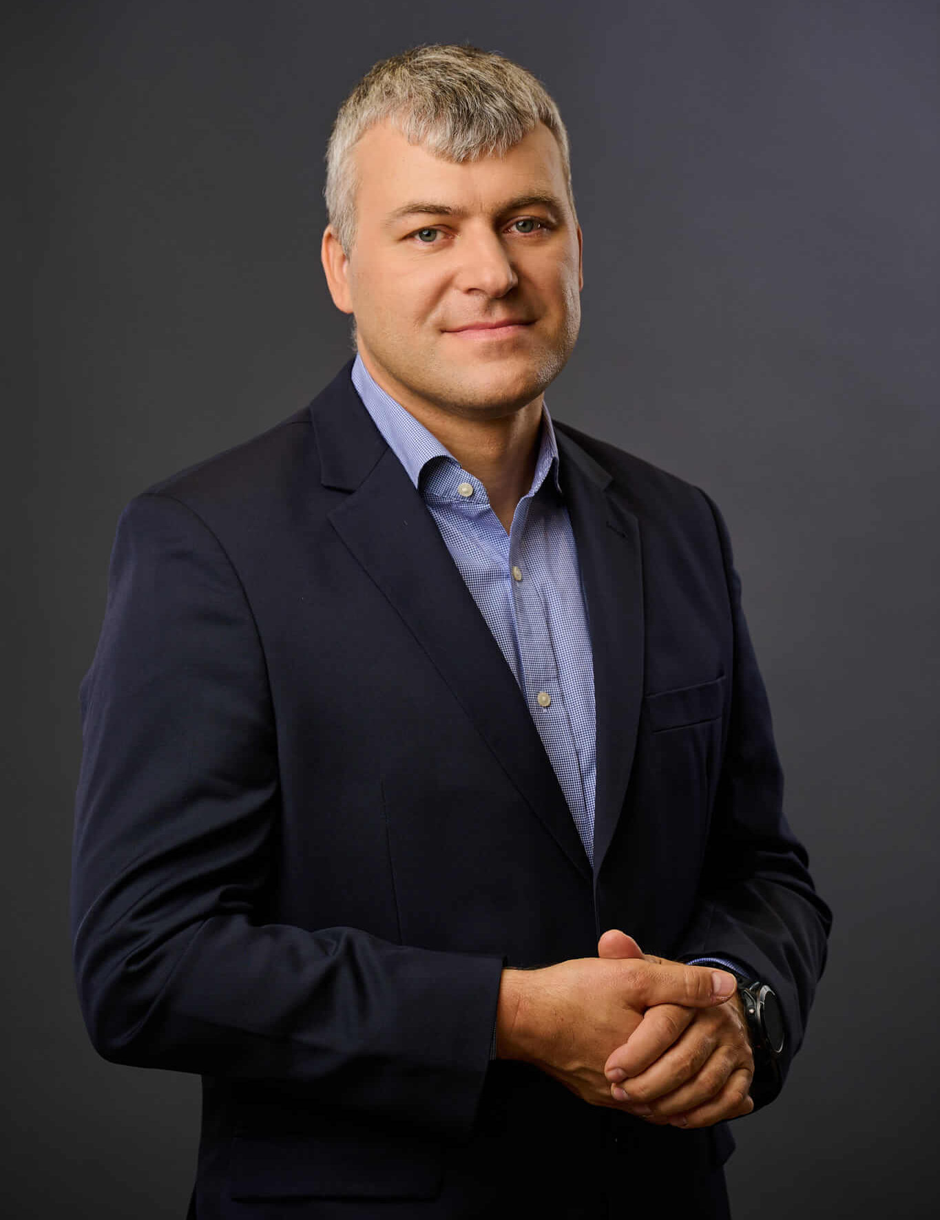 A man with short gray hair wearing a dark suit and light blue shirt stands against a plain dark background, looking at the camera with a slight smile and hands clasped in front of him.