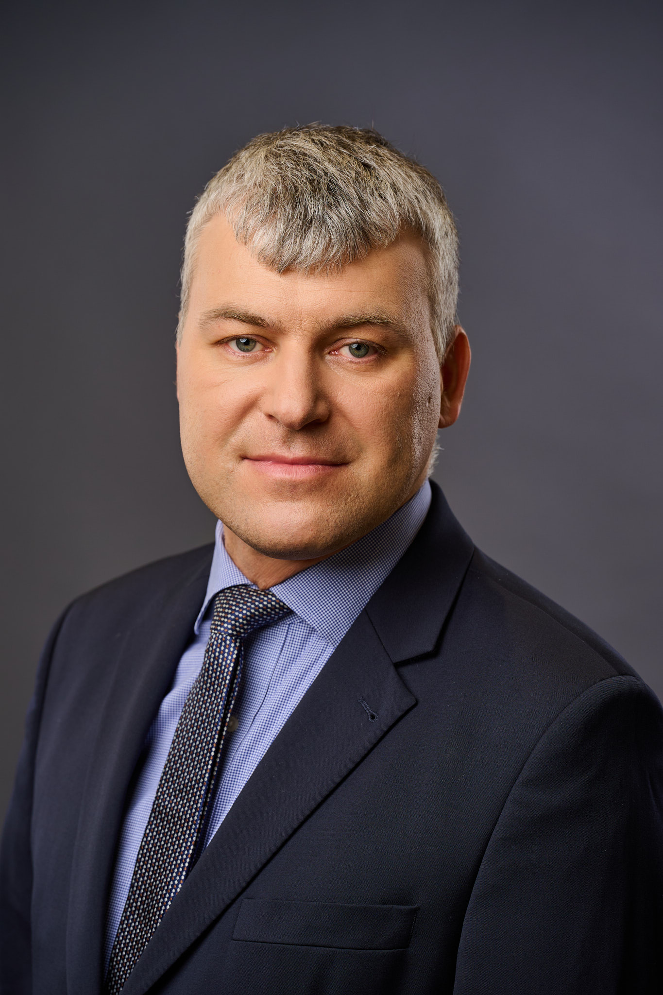 A man in a navy suit, light blue shirt, and patterned tie poses for a professional headshot against a plain dark background. He has short, light gray hair and a slight smile.