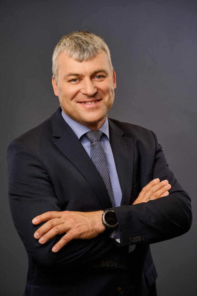 A man in a dark suit and tie stands with arms crossed, smiling confidently against a plain dark background. He has short gray hair and wears a watch on his left wrist.
