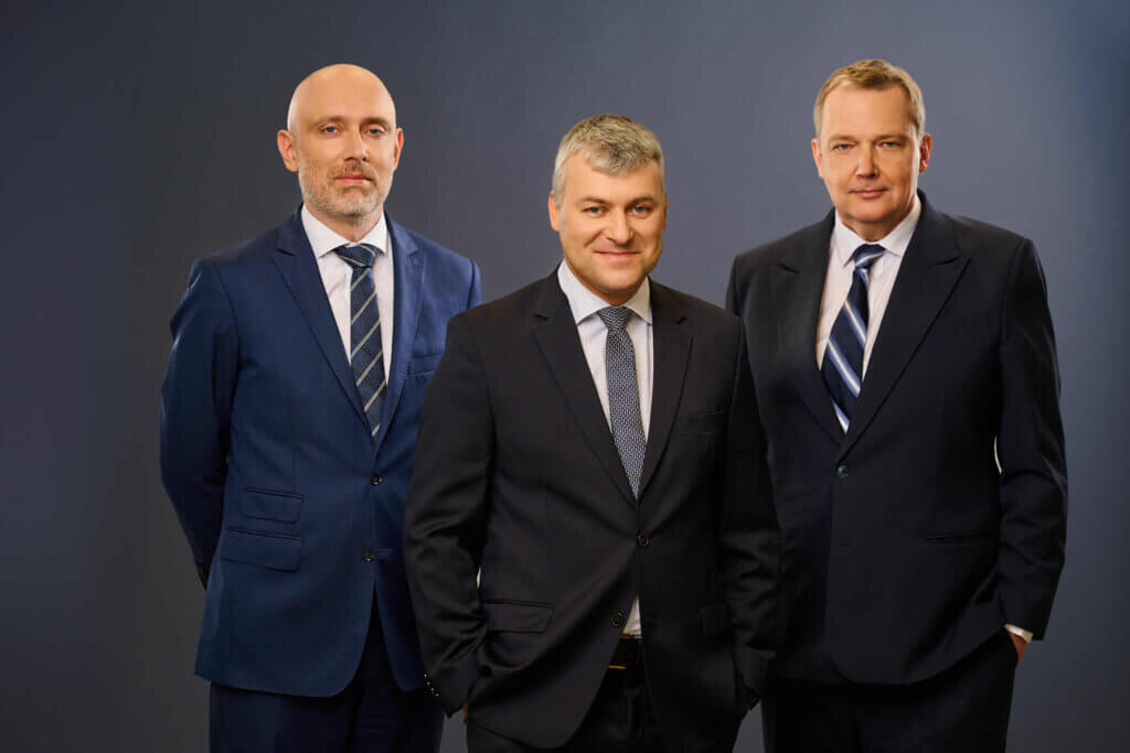 Three men in suits pose together against a plain, dark background. They are standing close, facing the camera, and all are dressed formally in business attire with ties.