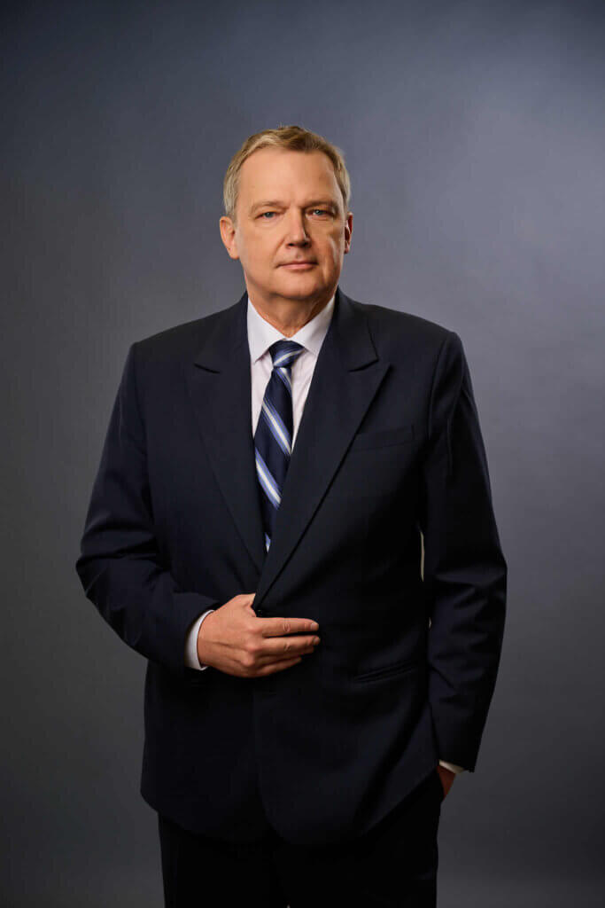 A middle-aged man in a dark suit and striped tie stands against a gray studio background, with one hand holding his jacket and the other in his pocket, looking confidently at the camera.