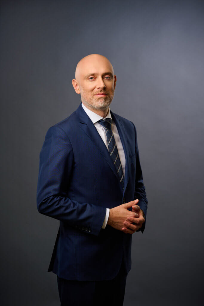 A bald man with a trimmed beard wearing a blue suit, white shirt, and striped tie stands against a plain gray background, posing with his hands clasped and a slight smile.