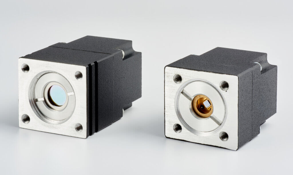 Two black and silver electronic components with square metal faces and circular centers, each featuring four corner screws, are placed side by side on a plain, light background.