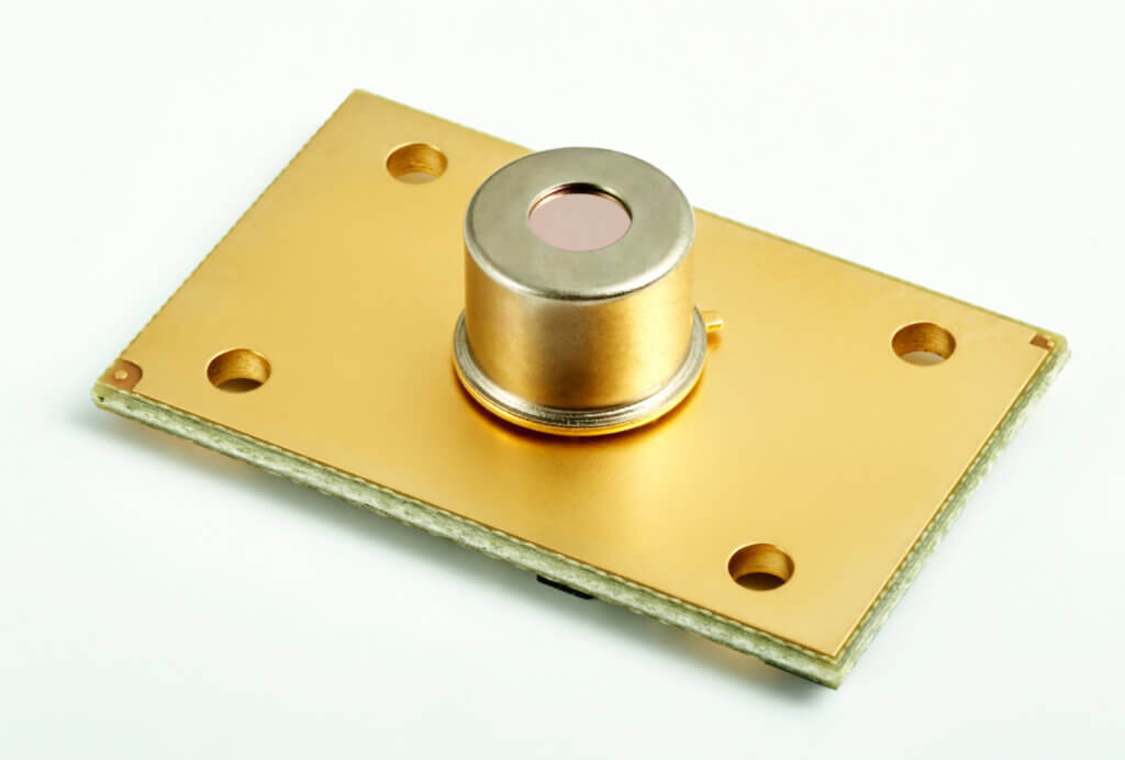 A gold-colored electronic sensor with a cylindrical component mounted on a rectangular base plate featuring four corner holes, placed on a white background.