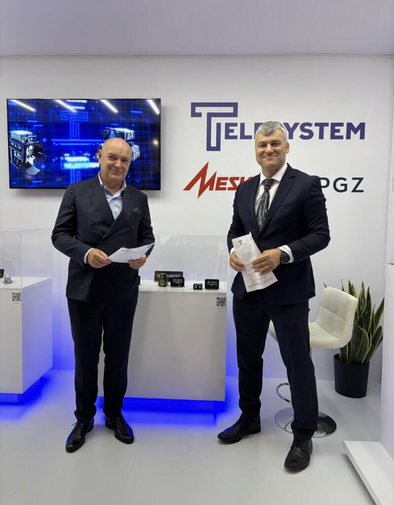Two men in suits stand smiling in a modern exhibition booth with Teldat, Telesystem, and Mesko PGZ logos on the wall behind them. One holds papers; tech equipment is displayed on a counter beside them.