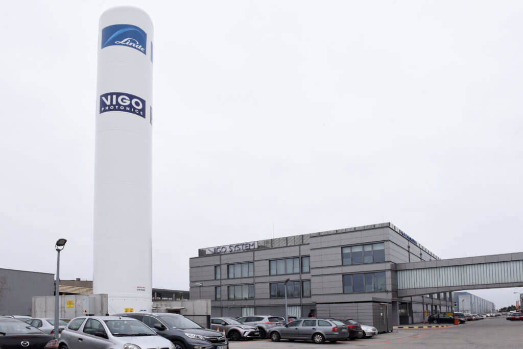 A large white industrial tower labeled Linde and Vigo Photonics stands beside a modern gray building complex with several parked cars and a skybridge on a cloudy day.