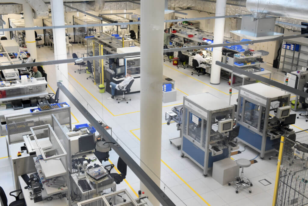 A clean, modern factory floor with automated machinery, workstations, and equipment arranged in organized rows. Few workers are visible, and the area is brightly lit with white walls and floors.