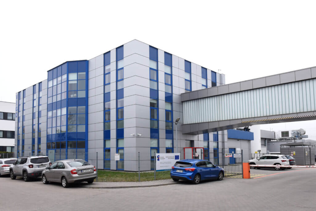 A modern, multi-story office building with blue-tinted windows and a skywalk connecting to another structure. Several parked cars are in the foreground, and the area appears clean and organized.
