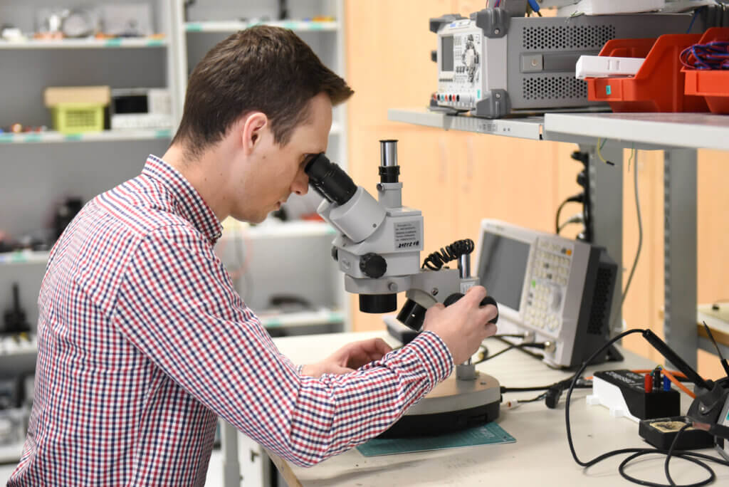 A person wearing a red and white checkered shirt is looking into a microscope at a workbench in a laboratory filled with electronic equipment and tools.