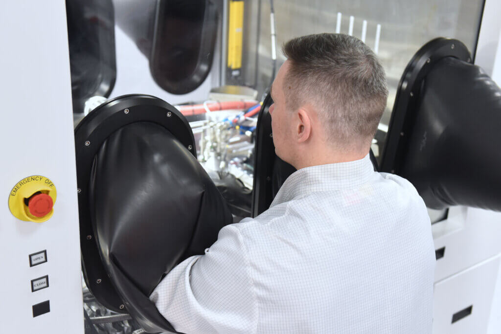 A person wearing a lab coat operates equipment inside a containment glove box, using black gloves attached to the enclosure. An emergency stop button is visible on the machine.