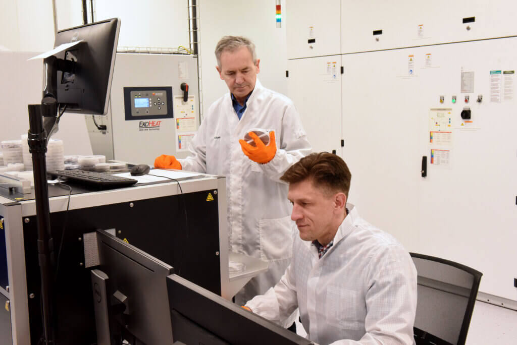 Two men in lab coats work in a laboratory. One is seated at a computer, focused on the screen, while the other stands nearby, wearing orange gloves and gesturing as he observes the workstation. Laboratory equipment surrounds them.