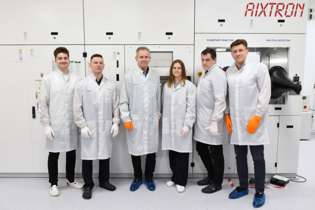 Six people wearing white lab coats and gloves stand in a row in front of large laboratory equipment labeled AIXTRON. They are posing for a group photo in a clean, modern laboratory setting.