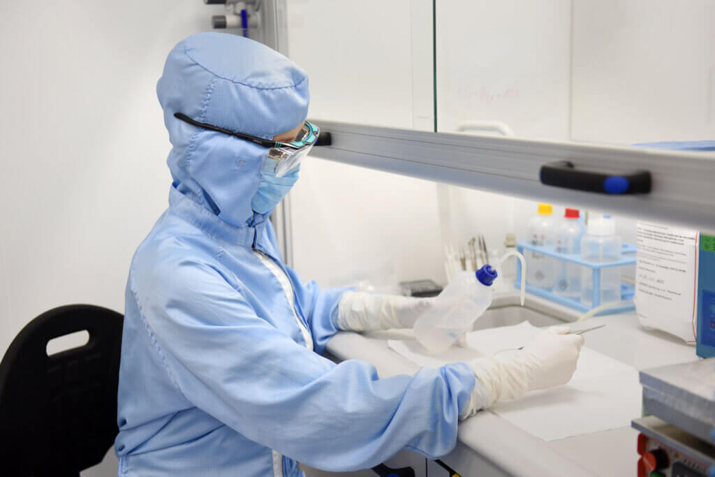 A scientist in full protective gear, including a mask, gloves, and goggles, works in a laboratory cleanroom, holding a plastic bottle and using lab equipment on a sterile work surface.