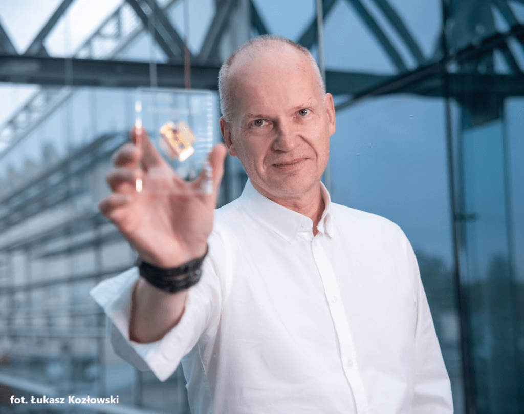 A man in a white shirt holds up a small transparent box containing a gold object, standing in front of a glass building. Photo credit: Łukasz Kozłowski.