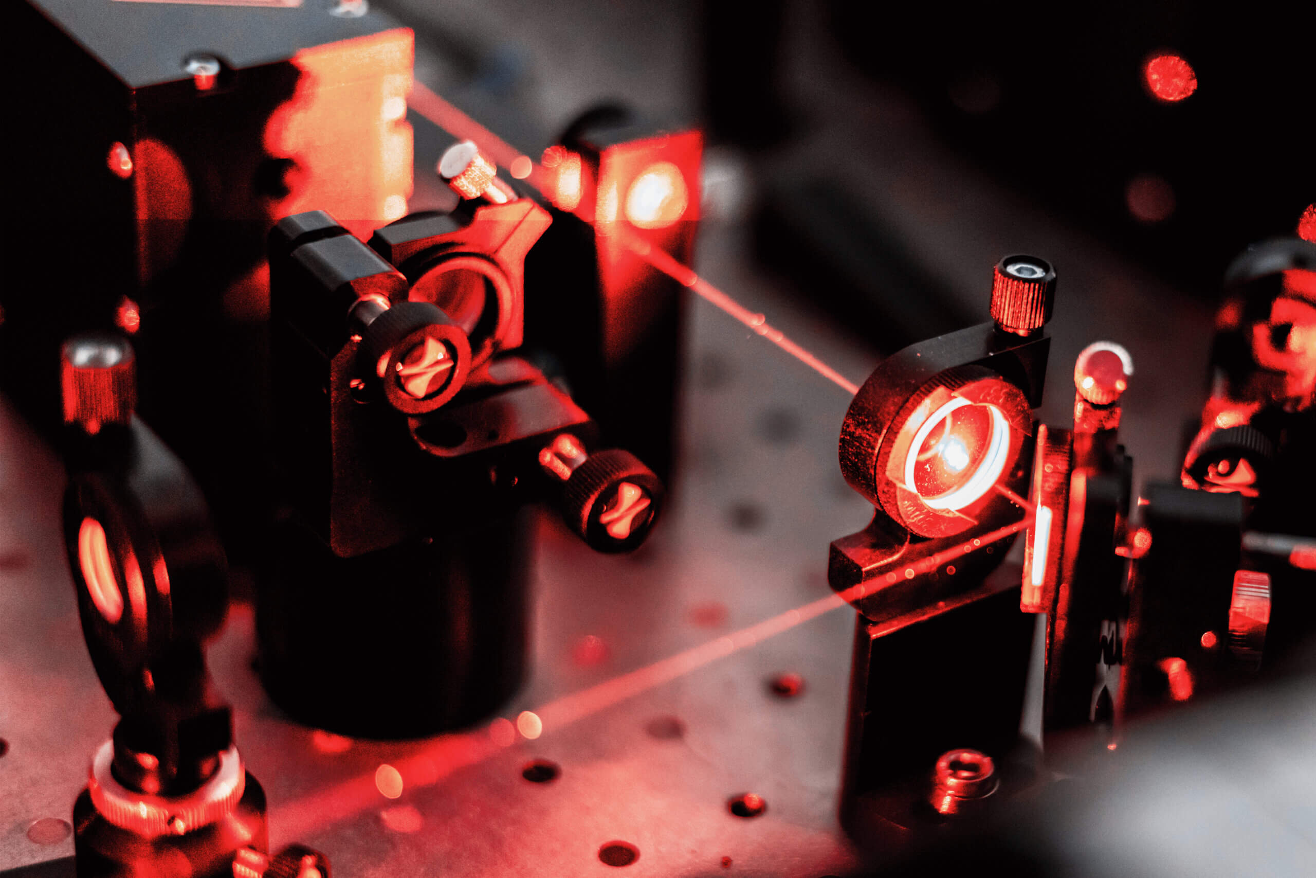 A close-up of a scientific optical setup with red laser beams passing through and reflecting off various lenses and mirrors mounted on a perforated metal surface.