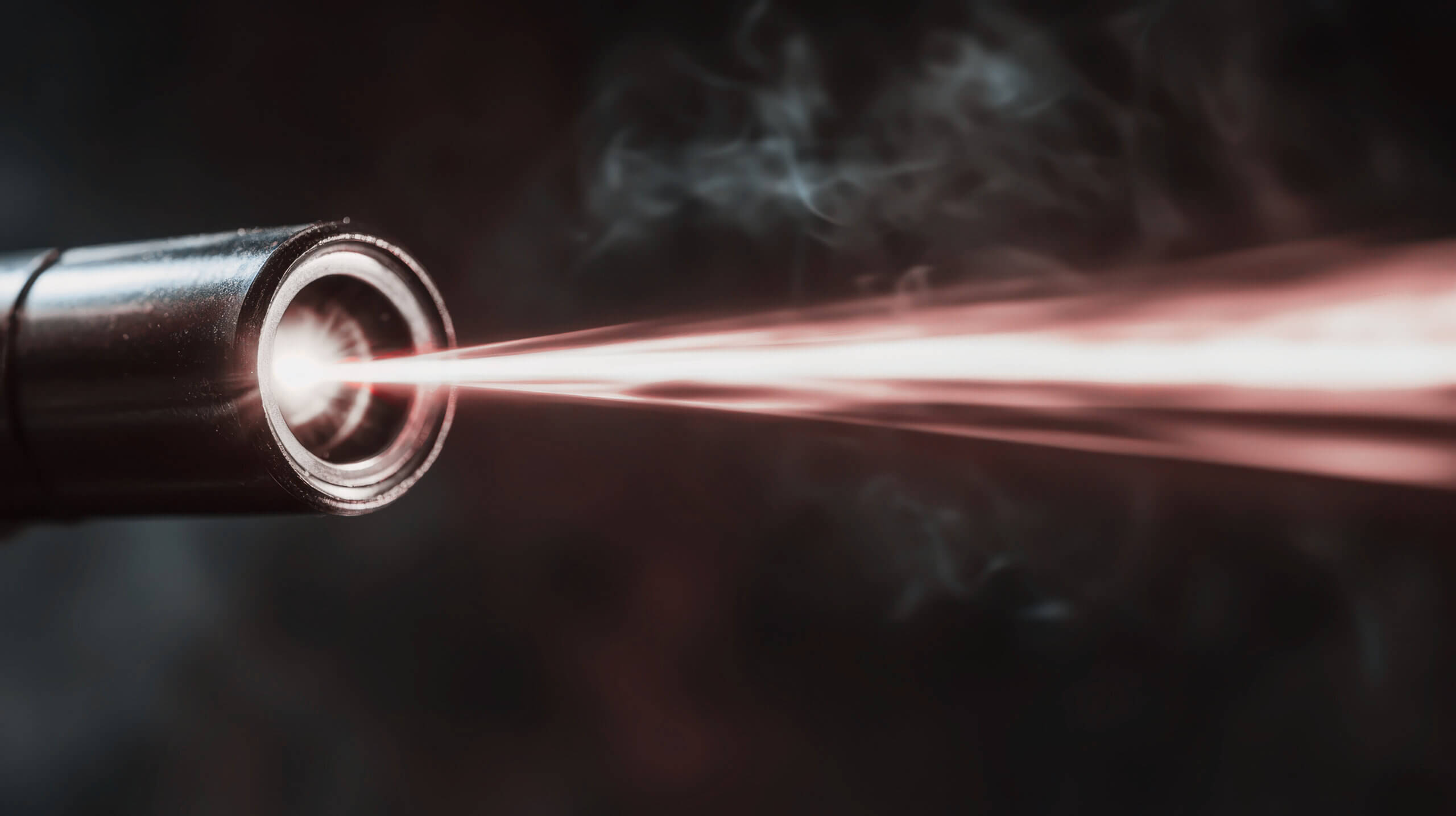 A close-up of a laser beam emitting from a metallic device, with red light cutting through smoke and a dark background.