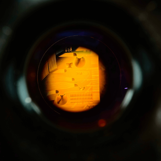 Close-up view through a circular lens, showing a detailed, illuminated microchip or electronic circuit with intricate lines and components, surrounded by a dark, blurred border.