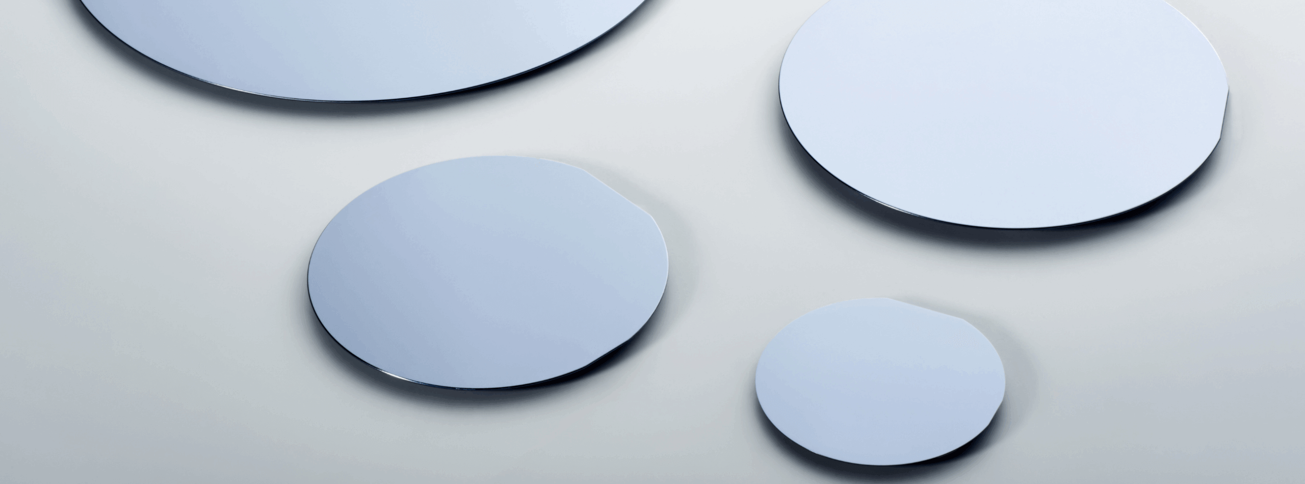 Four round, flat, reflective surfaces resembling mirrors or glass discs are arranged on a smooth, light grey background. The discs vary in size and have a clean, minimalist appearance.