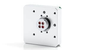 A square metallic device with rounded edges, multiple drilled holes, and a central cylindrical part featuring four red circular openings, set against a white background.