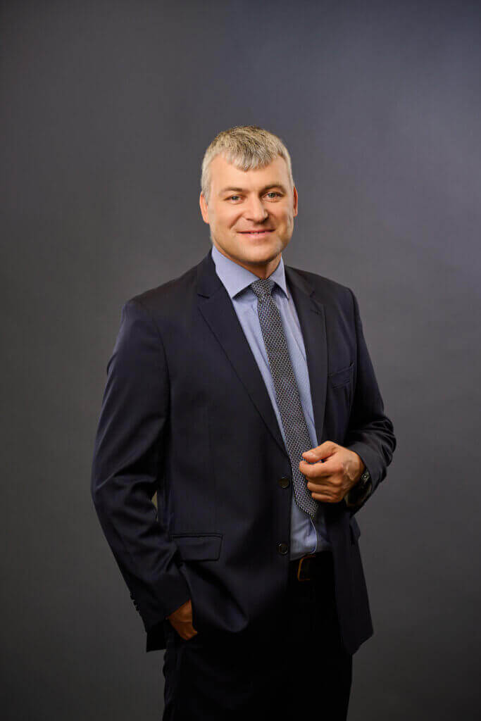 A man in a dark suit and blue patterned tie stands against a plain gray background, smiling with one hand in his pocket and the other adjusting his jacket.