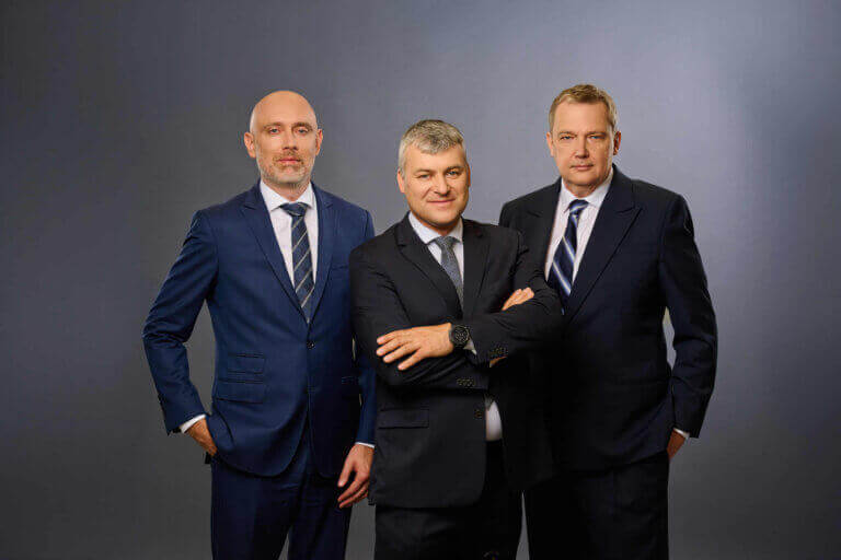 Three men in business suits pose against a plain gray background. Two stand on either side while the middle man stands slightly forward with arms crossed, all looking confidently at the camera.