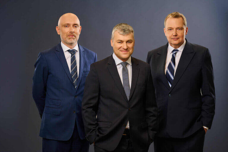 Three men in business suits stand side by side against a plain, dark background, facing the camera with neutral to slight smiles.