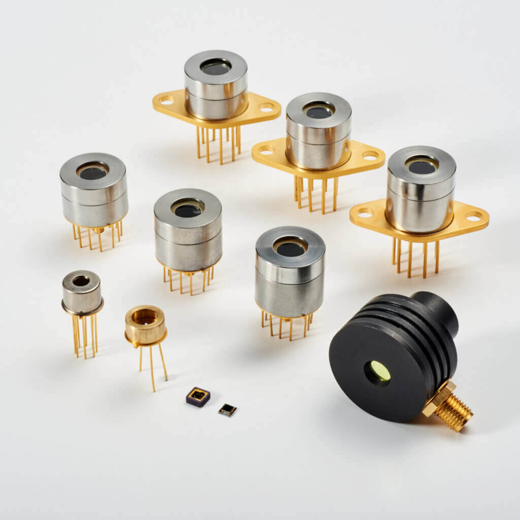 A variety of circular and rectangular electronic components, including metal and gold-plated cylindrical sensors with wire leads, and small square microchips, displayed on a white surface.