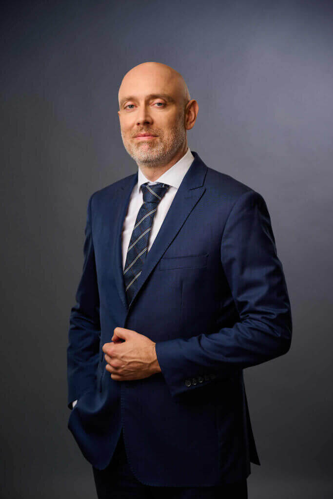 A bald man with a short beard, wearing a navy blue suit, white shirt, and striped tie, stands confidently against a dark gray background.