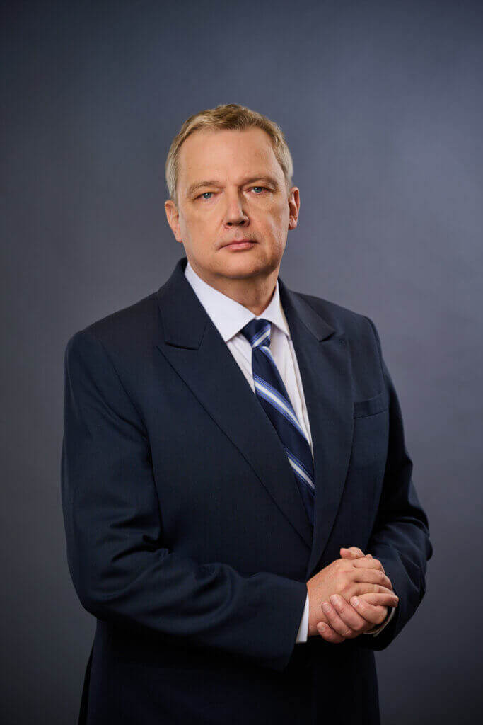 A middle-aged man with short blond hair wears a dark suit, white shirt, and striped tie, standing against a plain gray background with his hands clasped in front of him.