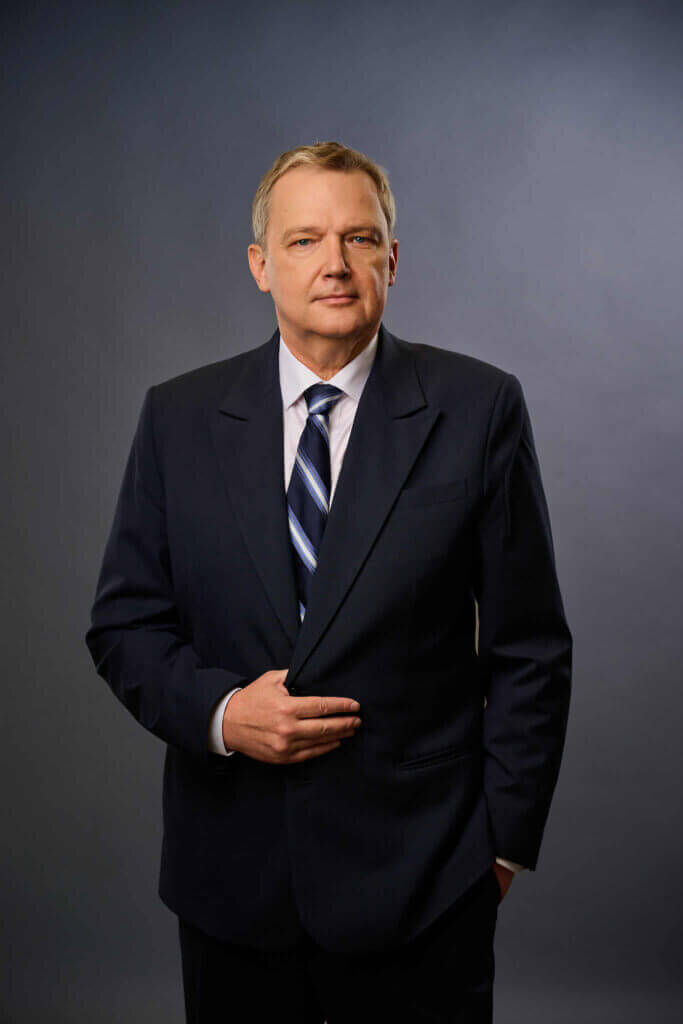 A middle-aged man in a dark suit and striped tie stands against a plain, dark gray background, looking confidently at the camera with one hand in his pocket and the other adjusting his jacket.