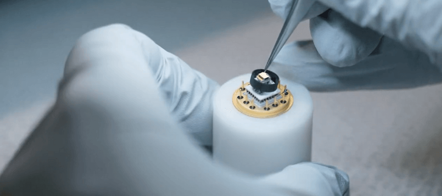 A gloved hand uses tweezers to carefully adjust a small electronic component on top of a cylindrical device, highlighting precision work in a laboratory or cleanroom setting.