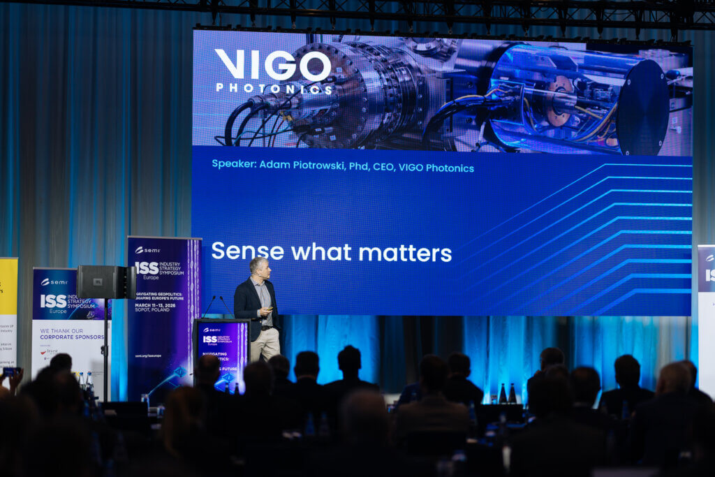 A man stands on stage giving a presentation titled “Sense what matters” for VIGO Photonics, with a large screen showing scientific equipment and the name Adam Piotrowski, PhD, CEO. The audience is seated in front.