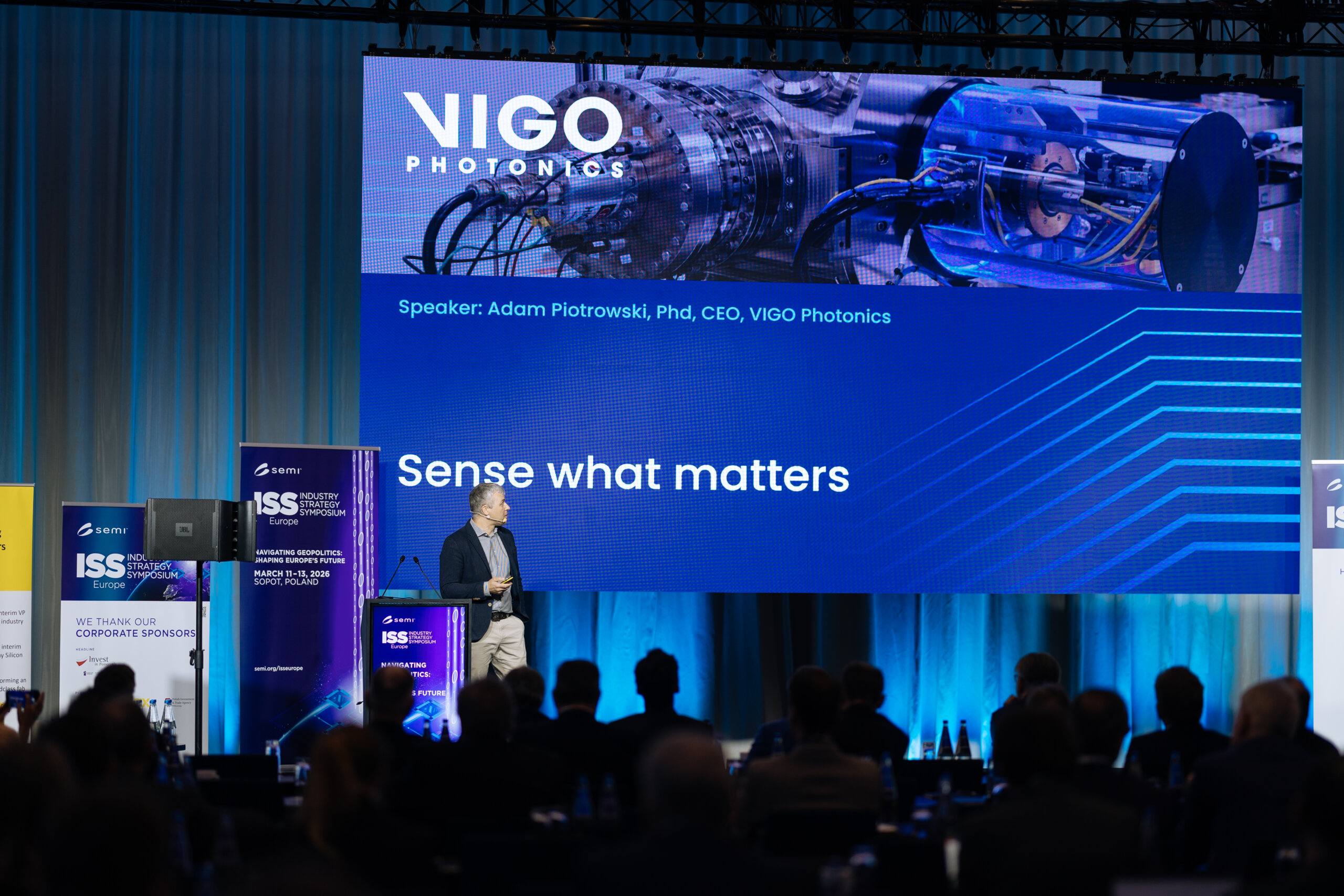 A man stands on stage giving a presentation titled “Sense what matters” for VIGO Photonics, with a large screen showing scientific equipment and the name Adam Piotrowski, PhD, CEO. The audience is seated in front.