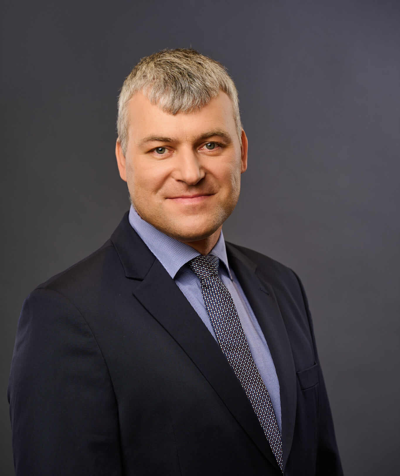 A man with short gray hair, wearing a dark suit, blue shirt, and patterned tie, stands in front of a plain gray background, smiling slightly at the camera.