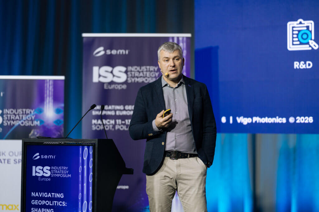 A man in a suit stands on stage giving a presentation at the ISS Industry Strategy Symposium Europe. He holds a device and stands near a podium, with banners and a projector screen in the background.
