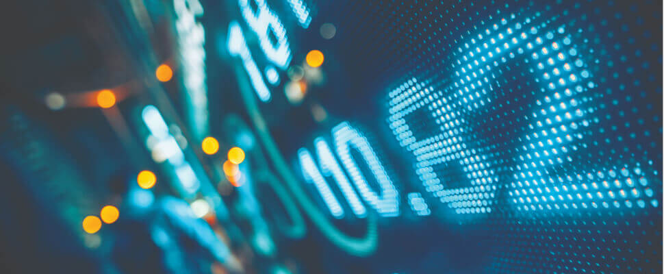 Close-up of a digital stock market display showing numbers and financial data in blue LED lights, with blurred orange lights in the background.