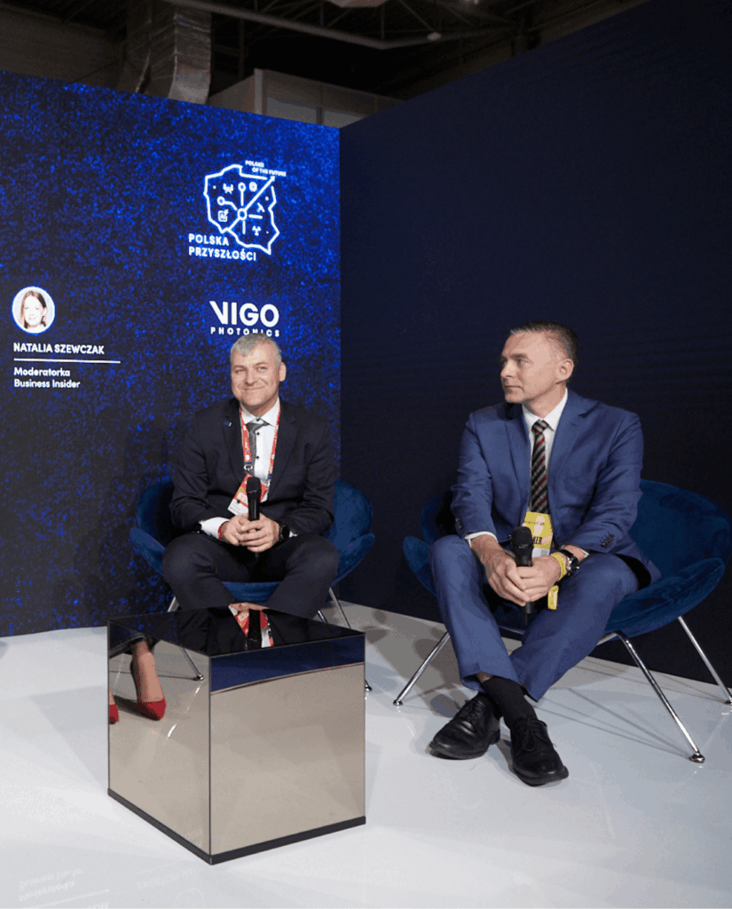 Two men in suits sit in blue chairs, on a stage with a dark blue backdrop featuring a Polska Przyszłości logo and Vigo Photonics branding. A reflective cube sits between them.