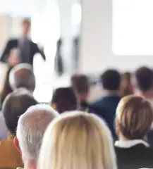 A blurred image of people seated, facing a presenter in a room. The focus is on the audience's heads, with the speaker and a screen in the background out of focus. The environment suggests a conference or seminar setting.