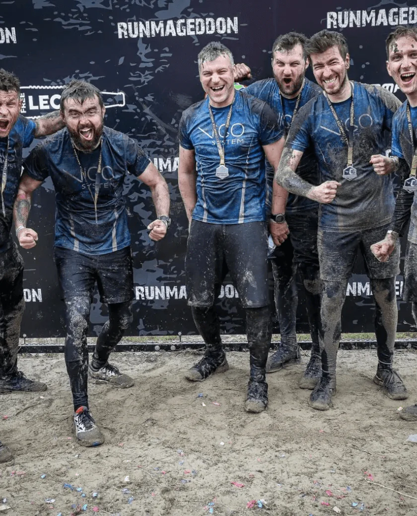 A group of six muddy participants in blue shirts and medals celebrate triumphantly after completing an obstacle race, standing on a sandy surface against a large "Runmageddon" backdrop. Some are cheering, showcasing their medals.