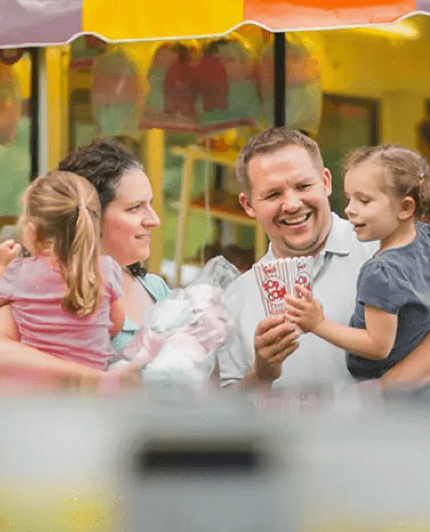 A happy family of four stands outdoors. The parents, a man and woman, are smiling and holding two young children. The background is colorful and festive, suggesting a fun setting.